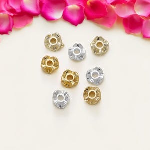 Può includere: Una collezione di nove perline a forma di fiore in tonalità oro e argento. Le perline hanno un bordo strutturato e increspato e un foro centrale. Petali di rosa rosa sparsi.