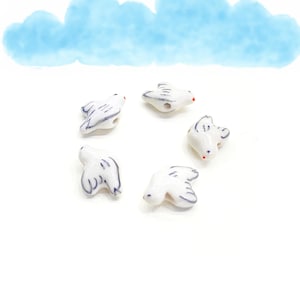 Peut inclure: Cinq perles en céramique blanche en forme de colombe avec des détails peints en bleu. Les colombes sont disposées en cercle sur un fond blanc.