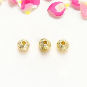 May include: Three gold-colored beads with intricate spiral patterns and a central hole. The beads are round and have a slightly textured surface. The background is white with pink and yellow flower petals.