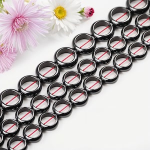 May include: A strand of black hematite beads with red accents. The beads are round and have a hole in the center. The beads are arranged in a row on a white surface.