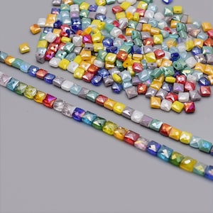 May include: A collection of colorful, square-shaped beads in various colors, including red, yellow, blue, and green. Some beads are strung into lines, while others are scattered. The beads have a shiny, iridescent finish.
