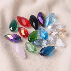 Pode incluir: Uma coleção de contas facetadas em forma de gota em várias cores, incluindo verde, vermelho, preto e iridescente. As contas estão dispostas sobre um tecido macio, branco-sujo, mostrando o seu corte e clareza.