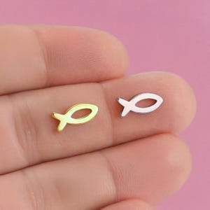 Könnte beinhalten: Zwei kleine Ohrringe in Fischform. Einer ist goldfarben mit hellgelbem Innenraum, der andere silberfarben. Die Ohrringe werden vor einem rosa Hintergrund präsentiert.