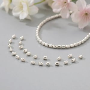 May include: A silver beaded necklace and scattered beads on a gray surface. The necklace is composed of small, square-shaped beads. Pink and white flowers are in the background.