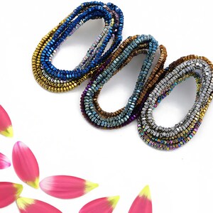 May include: Three strands of iridescent square beads in blue, brown, and silver. The beads are arranged in a circle and are on a white background.