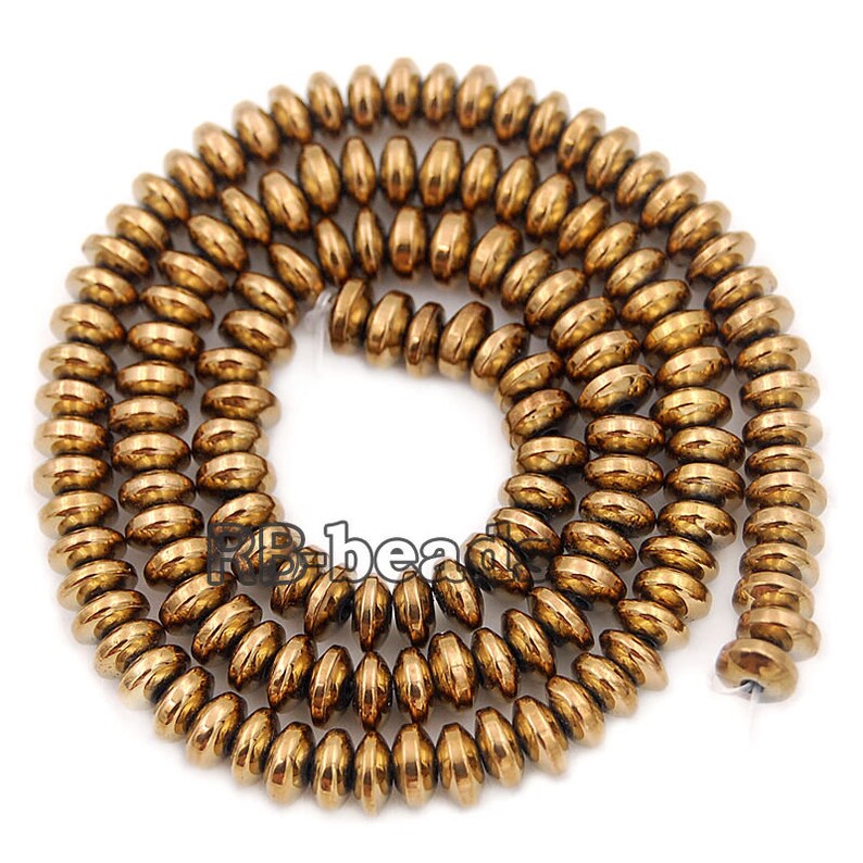 May include: A strand of gold-toned hematite beads, each bead is a flat, oval shape with a slightly rounded edge. The beads are strung together to form a long necklace.