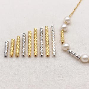 以下が含まれることがあります： テクスチャ加工されたツイストデザインの、銀色と金色をした金属製のチューブビーズの詰め合わせ。ビーズの長さは様々で、一列に並んでいます。パールと似たようなビーズのネックレスも写っています。