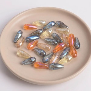 May include: A selection of iridescent, teardrop-shaped beads in orange, yellow, and blue-grey hues. The beads are arranged on a light beige plate, highlighting their faceted surfaces and reflective properties. Suitable for jewellery making.