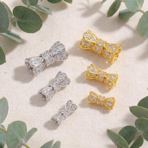 Könnte beinhalten: Eine Auswahl an schmuckartigen Schleifen. Einige sind silberfarben und mit kleinen, klaren Steinen besetzt. Andere sind goldfarben und ähnlich verziert. Der Schmuck wird auf einer neutralen, strukturierten Oberfläche mit grünen Blättern präsentiert.