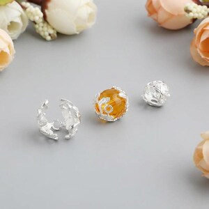 May include: Three silver-colored jewelry components. One is a floral-shaped clasp, and the other two are spherical beads. One bead has an orange-yellow center, and the other is solid silver. The items are on a gray surface with floral accents.