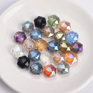 Può includere: Una collezione di perline esagonali sfaccettate in vari colori, tra cui nero, viola e blu. Le perline hanno una qualità iridescente e sono disposte su un piatto bianco.