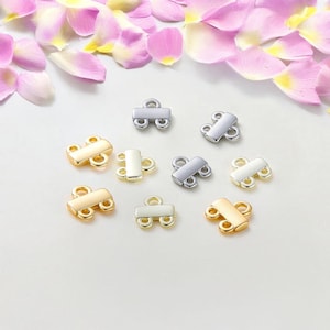 May include: A collection of metal jewelry clasps in gold, silver, and white gold tones. The clasps have a rectangular bar with two loops on each end. The background features pink and yellow flower petals.