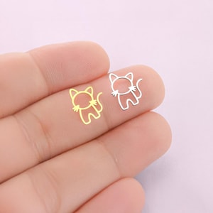 May include: Two small, cat-shaped charms. One is gold-colored, the other is silver-colored. The charms are cut-out designs, showing the outline of a cat with a tail and ears. The charms are held in a person's hand.