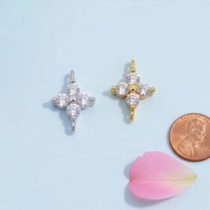 Puede incluir: Dos colgantes en forma de cruz, uno plateado y otro dorado, cada uno adornado con piedras preciosas redondas y transparentes. Una moneda de un centavo y un pétalo rosa también están en la imagen, sobre un fondo azul claro. Los colgantes son probablemente joyas.