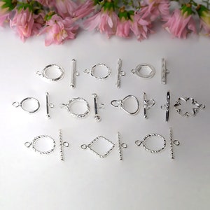 May include: A collection of 12 silver toggle clasps in various shapes and designs, including round, oval, square, and flower shapes. The clasps are arranged in rows on a white surface.