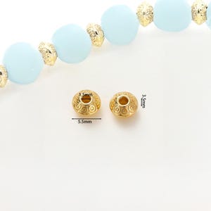 May include: Two gold-colored, decorative beads with a detailed swirl pattern. Each bead is 5.5mm wide and 3.5mm high, with a 1.5mm hole. The beads are shown with a strand of light blue beads and gold-colored spacers.