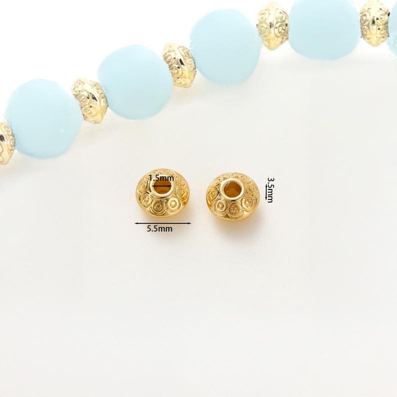 May include: Two gold-colored, decorative beads with a detailed swirl pattern. Each bead is 5.5mm wide and 3.5mm high, with a 1.5mm hole. The beads are shown with a strand of light blue beads and gold-colored spacers.