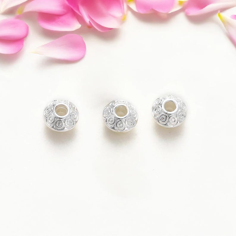 May include: Three silver-colored beads with a detailed spiral design. Each bead has a central hole and a rounded shape. Pink and white flower petals are scattered in the background, creating a soft, floral aesthetic.