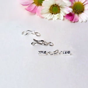May include: Three silver jewellery clasps are shown against a white backdrop, with pink and white flowers in the background. The clasps are of different designs, including a hook and eye style and a barrel clasp.