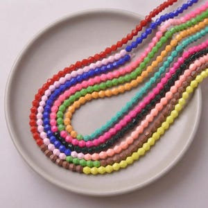 May include: A collection of colorful faceted beads in various shades, including red, blue, green, and pink, arranged on a light gray plate. The beads are strung together, creating a vibrant display of colors. Ideal for jewelry making or craft projects.