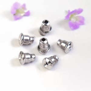 Op de afbeelding: Zes zilverkleurige oorbelruggen liggen op een witte ondergrond. De oorbelruggen hebben een cilindrische vorm met een afgerond uiteinde en een klein gaatje. Twee paarse bloemen zijn op de achtergrond.