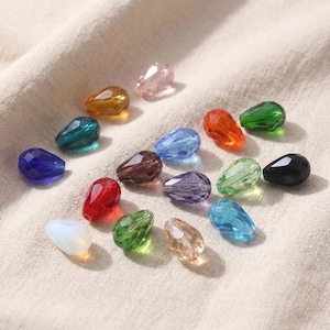May include: A collection of teardrop-shaped beads in various colors, including blue, green, red, and clear. The beads are faceted, reflecting light, and are arranged on a neutral-colored fabric. These beads are suitable for jewelry making and crafting.