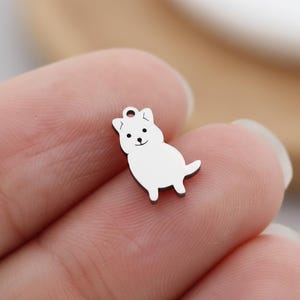 May include: A small, silver-colored charm in the shape of a dog. The charm has a loop at the top for attaching to a necklace or bracelet. The dog has a simple, cartoon-like design with black eyes and a smiling mouth.