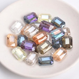 Könnte beinhalten: Eine Sammlung rechteckiger, facettierter Perlen in verschiedenen Farben, darunter klar, lila, orange und blau. Die Perlen sind auf einer weißen, flachen Schale angeordnet und scheinen aus Glas oder Kristall zu bestehen.