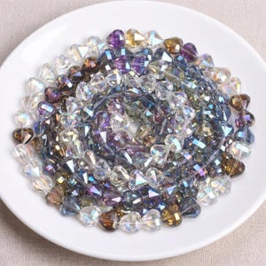 Peut inclure: Un ensemble de perles irisées en forme de cœur, dans des tons de violet, de marron et de transparent, disposées sur une assiette blanche. Les perles ont une surface à facettes, créant un effet scintillant. Les perles sont probablement destinées à la fabrication de bijoux.