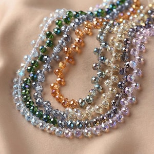 Pode incluir: Close-up de vários fios de colares de contas em várias cores, incluindo verde, laranja, prata e roxo. As contas são facetadas e parecem ser de cristal ou vidro, refletindo a luz. Os colares estão dispostos em um padrão circular.