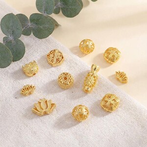 May include: A collection of gold-colored beads in various shapes, including spheres, lotus flowers, and geometric designs. The beads are displayed on a white fabric surface, with a sprig of green leaves in the background.