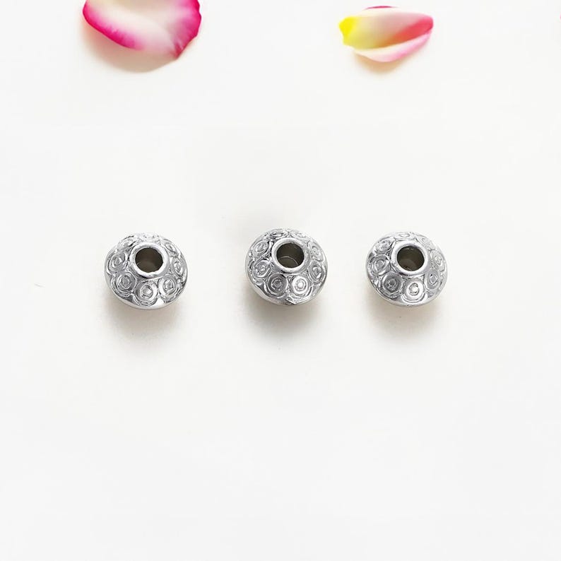 May include: Three silver-colored beads with a detailed floral pattern and a central hole. The beads are arranged on a white surface, with pink and yellow flower petals in the background. These beads are suitable for jewelry making.