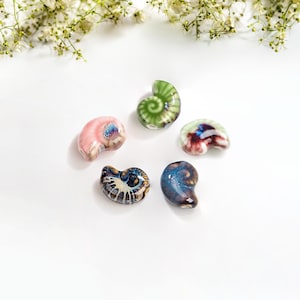 May include: Five ceramic seashell-shaped beads in various colors and patterns. The beads are green, pink, blue, and brown with white accents.
