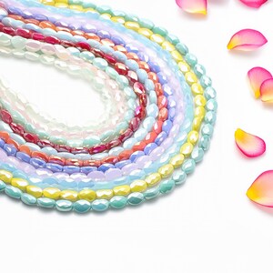 Puede incluir: Una colección de cuentas ovaladas de colores en varios tonos pastel, incluyendo blanco, rosa, rojo, azul y amarillo. Las cuentas están dispuestas en un patrón circular, con algunos pétalos de rosa dispersos en el fondo. Ideal para la fabricación de joyas.