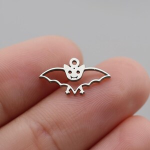 Op de afbeelding: Een kleine, zilverkleurige vleermuisbedel met een lachend gezicht en open vleugels. De bedel heeft een lus aan de bovenkant om aan sieraden of andere knutselwerkjes te bevestigen. De vleugels van de vleermuis hebben een geschulpte rand.