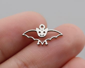 May include: A small, silver-colored bat charm with a smiling face and open wings. The charm has a loop at the top for attaching to jewelry or other crafts. The bat's wings have a scalloped edge.