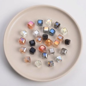 May include: A collection of iridescent, square-shaped beads in various colors, including orange, blue, and clear, are scattered on a beige plate. The beads have a faceted surface, reflecting light and creating a sparkling effect. These beads are ideal for jewelry making.
