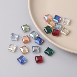 May include: A selection of square, iridescent glass beads in shades of blue, green, orange, and red. Some beads are on a beige plate, others are scattered on a grey surface. These beads are suitable for jewellery making and crafts.