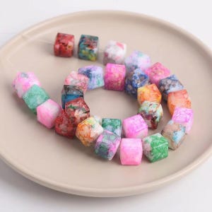 Puede incluir: Un collar de cuentas cúbicas multicolores, rosas, verdes, azules, naranjas y rojas. Las cuentas tienen un aspecto veteado y están ensartadas en un cordón transparente. Las cuentas se exhiben en un plato beige.