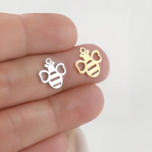 May include: Two bee-shaped charms, one silver and one gold, are displayed. The charms have a striped body, heart-shaped wings, and a crown. The silver charm is on the left, and the gold charm is on the right.