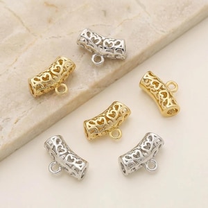 May include: An assortment of decorative metal tube beads. The beads are gold and silver, with a heart-shaped cutout design. Each bead has a small loop at one end. The beads are arranged on a light-colored surface.