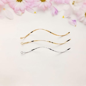 Puede incluir: Tres cadenas onduladas, una plateada y dos doradas, se colocan sobre una superficie blanca. Las cadenas tienen la misma longitud y un diseño simple y elegante.