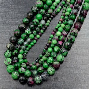 May include: A strand of green, black, and pink gemstone beads. The beads are round and have a smooth, polished finish. The beads are strung on a clear string.