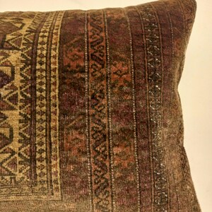 May include: Close-up of a rectangular pillow with a detailed, patterned design. The pillow features a mix of brown, tan, and burgundy colors. The design includes geometric shapes and vertical stripes, creating a textured appearance. The pillow is likely made of a woven fabric.