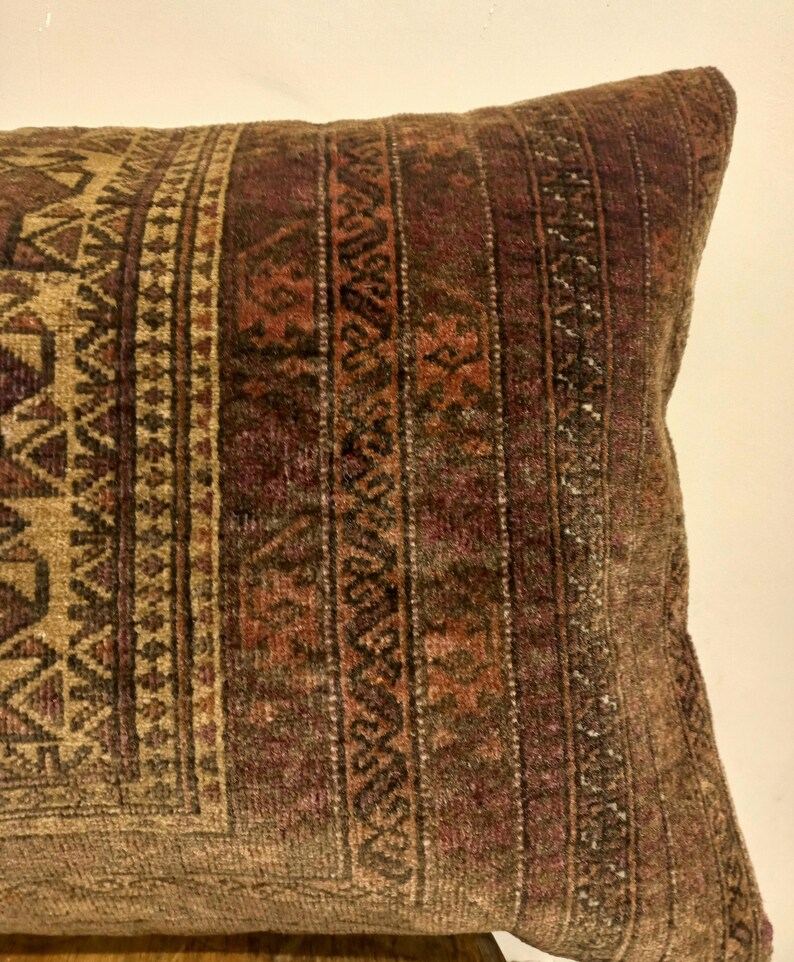 May include: Close-up of a rectangular pillow with a detailed, patterned design. The pillow features a mix of brown, tan, and burgundy colors. The design includes geometric shapes and vertical stripes, creating a textured appearance. The pillow is likely made of a woven fabric.