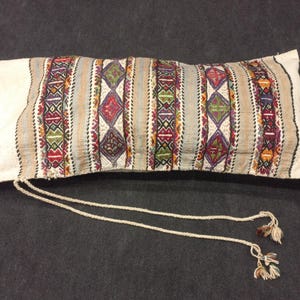 May include: A long, rectangular pillow with a white fabric cover and colorful embroidered stripes. The pillow has a drawstring closure and two tassels.