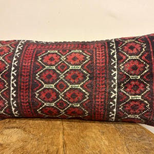 May include: Rectangular decorative pillow with a red, black, and white geometric pattern. The pillow has a central panel with repeating diamond shapes and borders with intricate designs. The pillow is placed on a wooden surface.