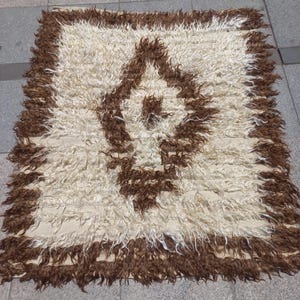 Peut inclure: Un tapis rectangulaire à poils longs et texturés, dans des tons crème et marron. Le motif présente une bordure marron épaisse et un losange central marron, contrastant avec un fond crème composé de fibres longues et ébouriffées.