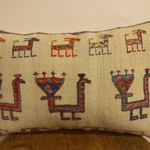 May include: Rectangular beige pillow with repeating geometric animal designs in red, orange, and blue. The pillow is made of a woven fabric and appears to be handmade. Decorative home decor item.