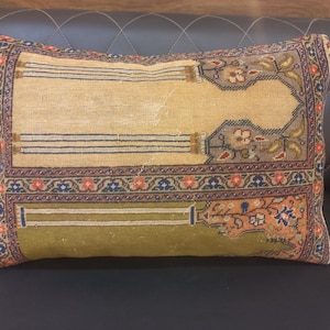 May include: Rectangular decorative pillow with a vintage rug design. The pillow features a beige center with blue, red, and green patterned borders and accents. The pillow is placed on a dark brown leather surface.